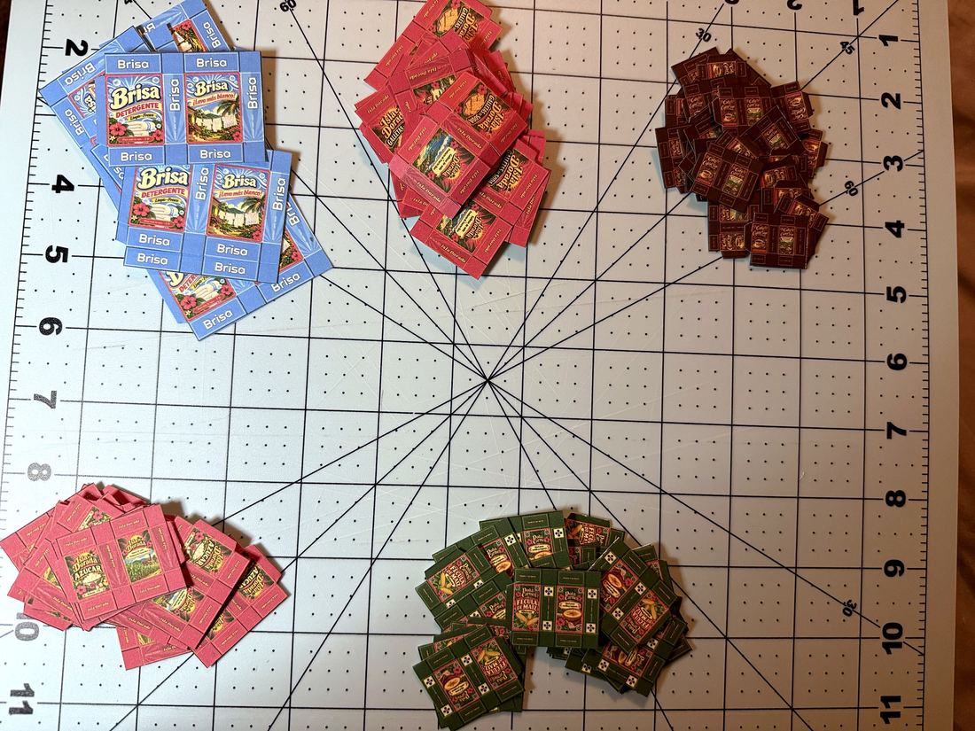 Piles of cut, unfolded box templates for detergent, crackers, sugar, coffee and cornstarch on a cutting mat